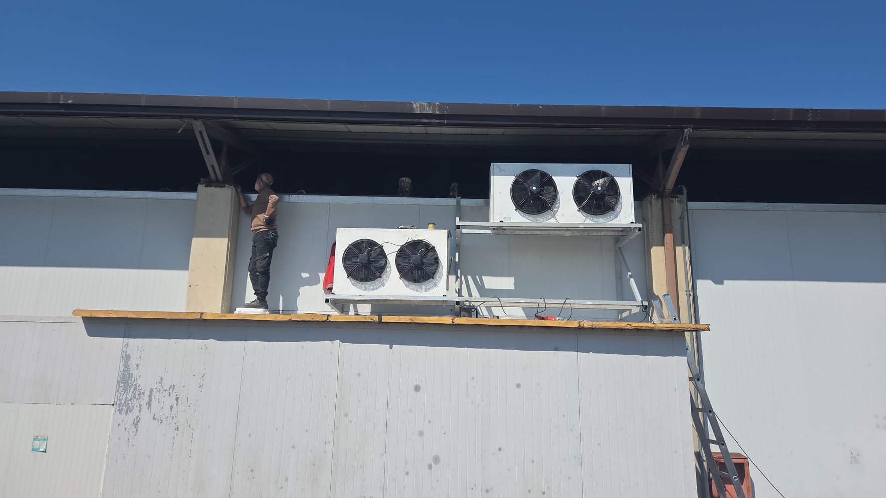 Tecnico su un tavolato di servizio mentre monta due aeroevaporatori sulla parete di un capannone
